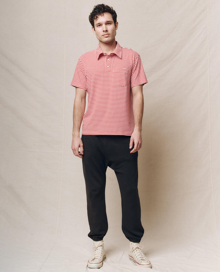 A person stands before a beige backdrop, wearing THE GREAT. SP25 POLO LOUNGE MEN's The Men's Lacrosse Polo in Sun Cherry Stripe, dark pants, and white sneakers, with a neutral expression.