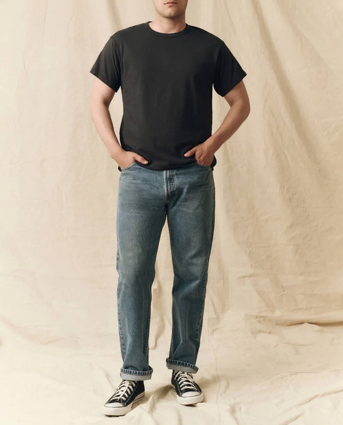 Against a beige fabric backdrop, a person wears THE GREAT. FALL 23 MEN The Men's Pure Knits Boxy Crew in Almost Black, paired with blue jeans and black-and-white sneakers, hands in pockets.