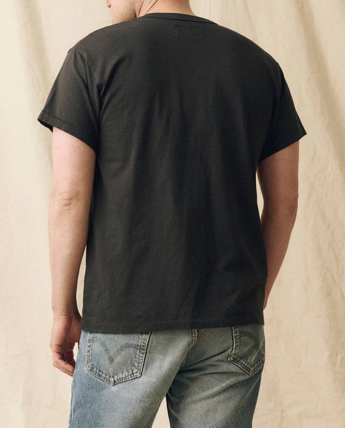 A person wearing THE GREAT. FALL 23 MEN The Men's Pure Knits Boxy Crew in Solid -- Almost Black and light blue jeans is shown from the back, standing against a neutral fabric background.
