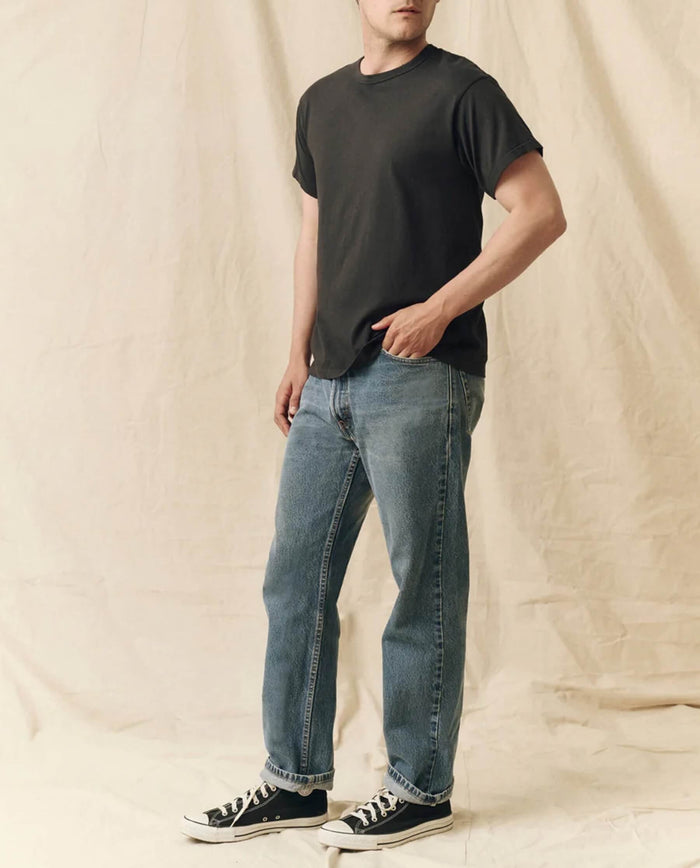 A person stands indoors against a beige fabric backdrop wearing THE GREAT. FALL 23 MEN The Men's Pure Knits Boxy Crew in Solid -- Almost Black, paired with blue jeans and black sneakers.