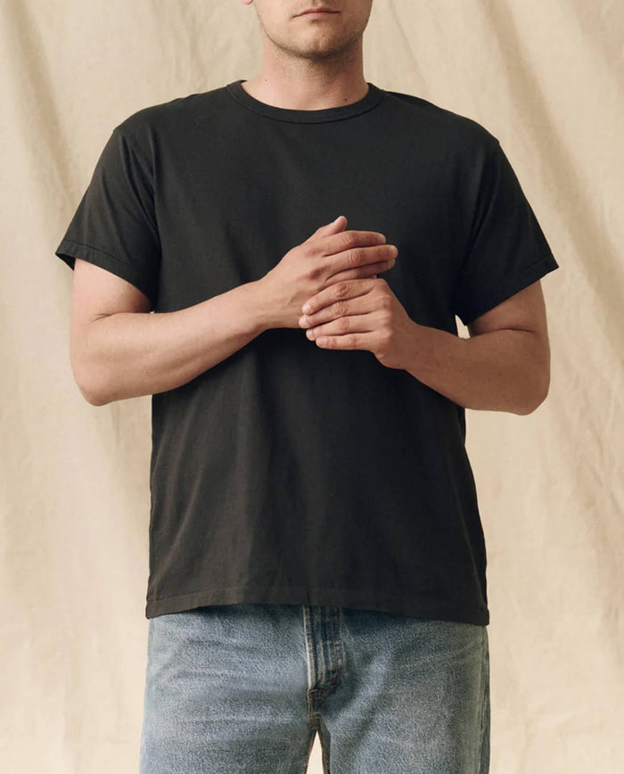 A person wears THE GREAT. FALL 23 MEN The Men's Pure Knits Boxy Crew, Solid -- Almost Black, paired with blue jeans, standing against a beige fabric background with hands clasped at chest level.