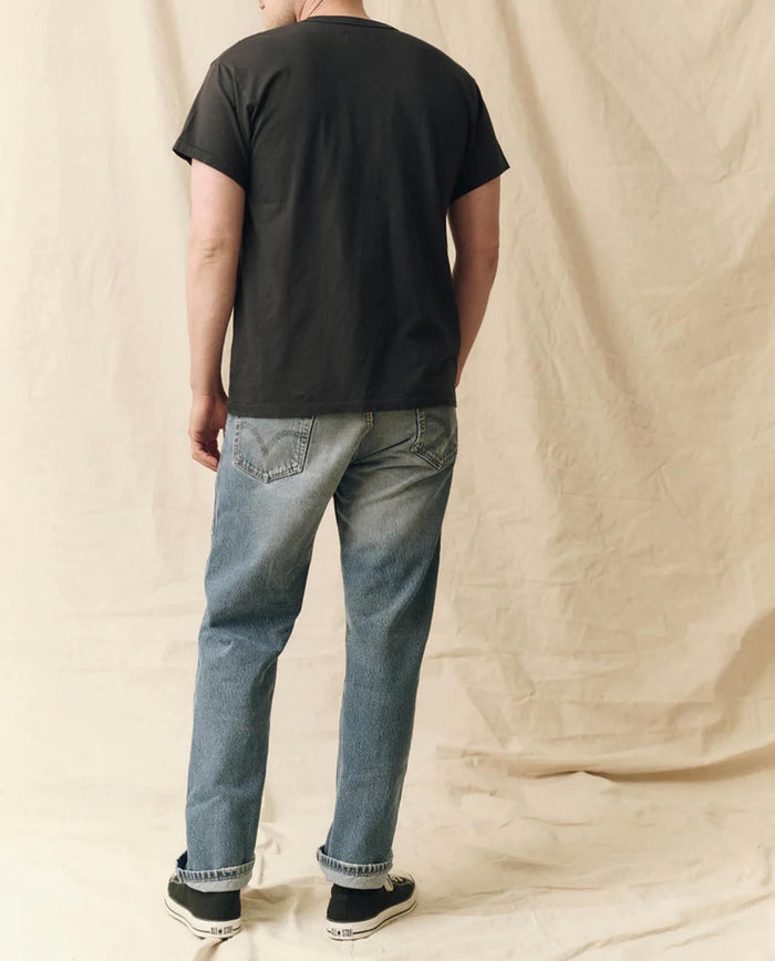 A person stands facing away, wearing THE GREAT. FALL 23 MEN The Men's Pure Knits Boxy Crew in Solid -- Almost Black, paired with blue jeans and black sneakers, against a beige fabric backdrop.