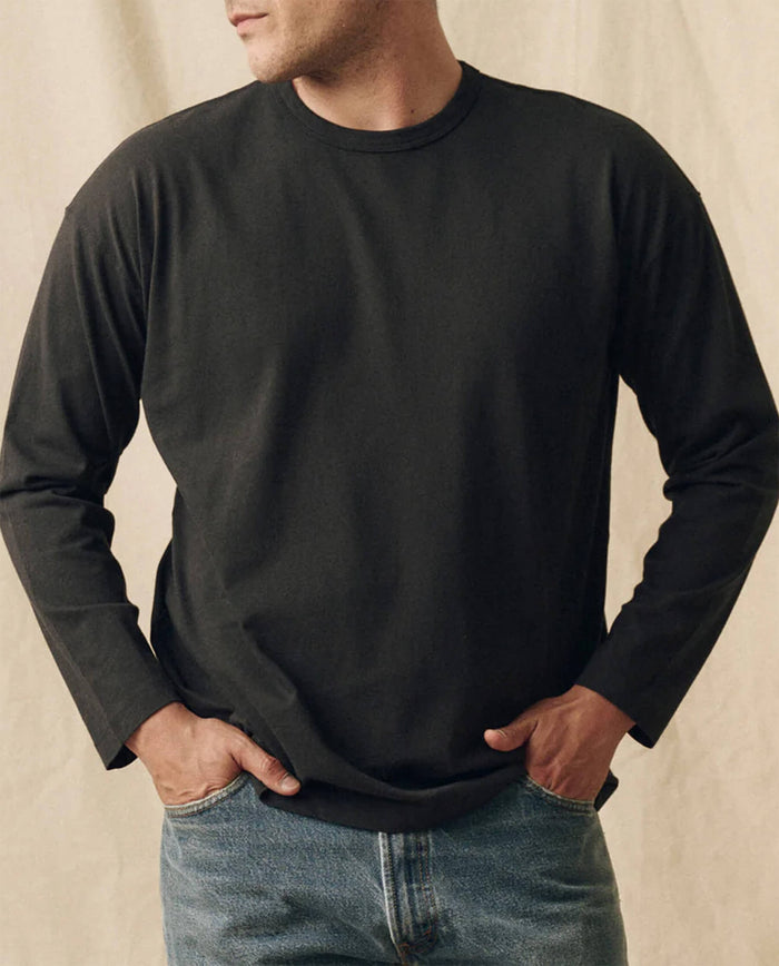 A person wears THE GREAT. FALL 23 MEN The Men's Pure Knits Long Sleeve Boxy Crew in Solid -- Almost Black with blue jeans, standing hands-in-pockets against a neutral background.