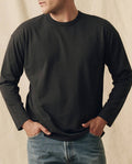 A person wears THE GREAT. FALL 23 MEN The Men's Pure Knits Long Sleeve Boxy Crew in Solid -- Almost Black with blue jeans, standing hands-in-pockets against a neutral background.