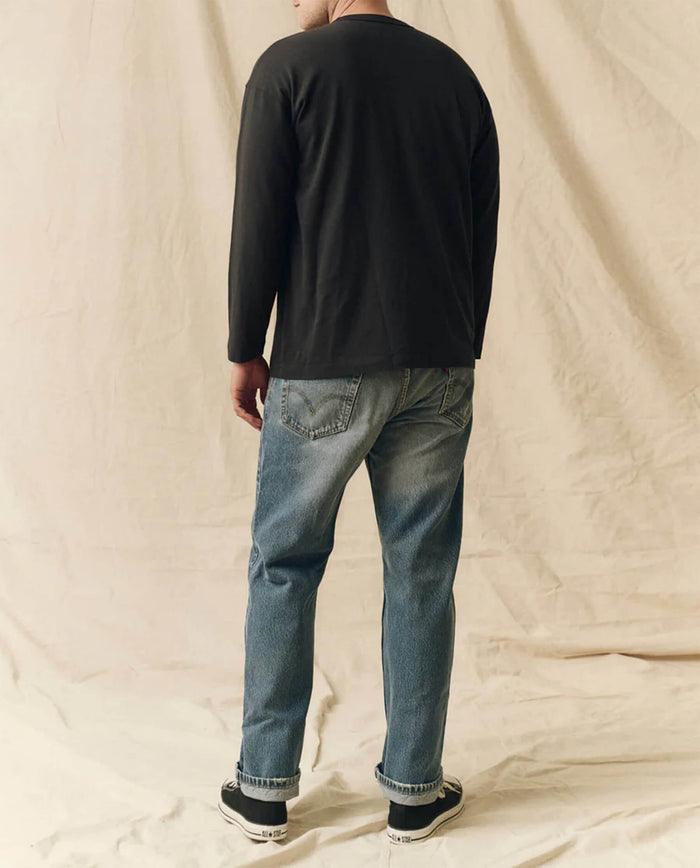 A person faces away from the camera wearing THE GREAT. FALL 23 MEN The Men's Pure Knits Long Sleeve Boxy Crew in Almost Black, blue jeans, and black-and-white sneakers against a neutral fabric backdrop.