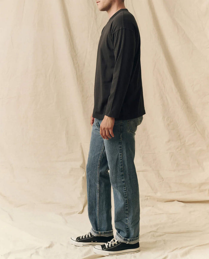 A person in profile wears THE GREAT. FALL 23 MEN The Men's Pure Knits Long Sleeve Boxy Crew in Almost Black, paired with blue jeans and black sneakers, standing before a beige fabric backdrop.