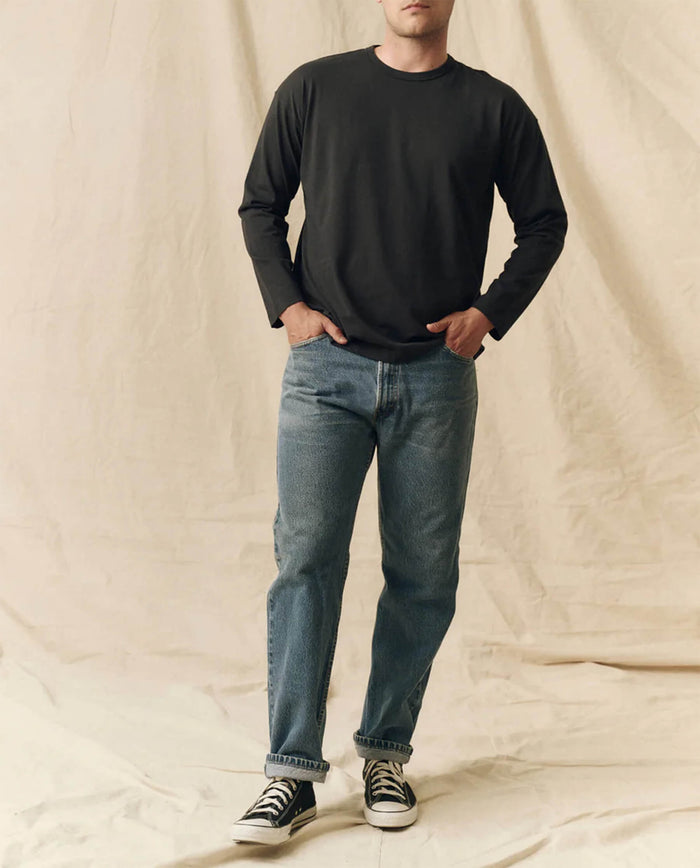 A person stands against a neutral fabric backdrop wearing THE GREAT. FALL 23 MEN's The Men's Pure Knits Long Sleeve Boxy Crew in Almost Black, paired with blue jeans and black-and-white sneakers.