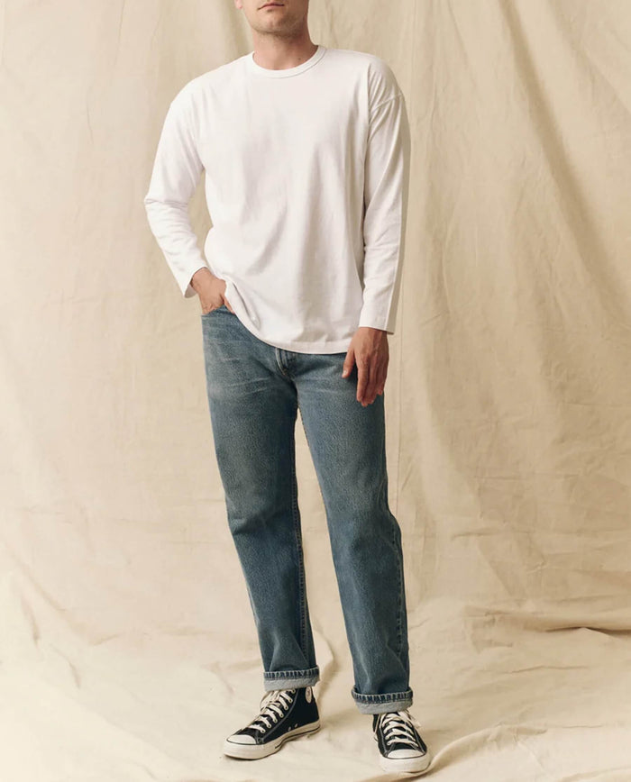 A person stands against a beige fabric backdrop, wearing THE GREAT. FALL 23 MEN's The Men's Pure Knits Long Sleeve Boxy Crew in True White, paired with blue jeans and black high-top sneakers.