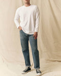 A person stands against a beige fabric backdrop, wearing THE GREAT. FALL 23 MEN's The Men's Pure Knits Long Sleeve Boxy Crew in True White, paired with blue jeans and black high-top sneakers.