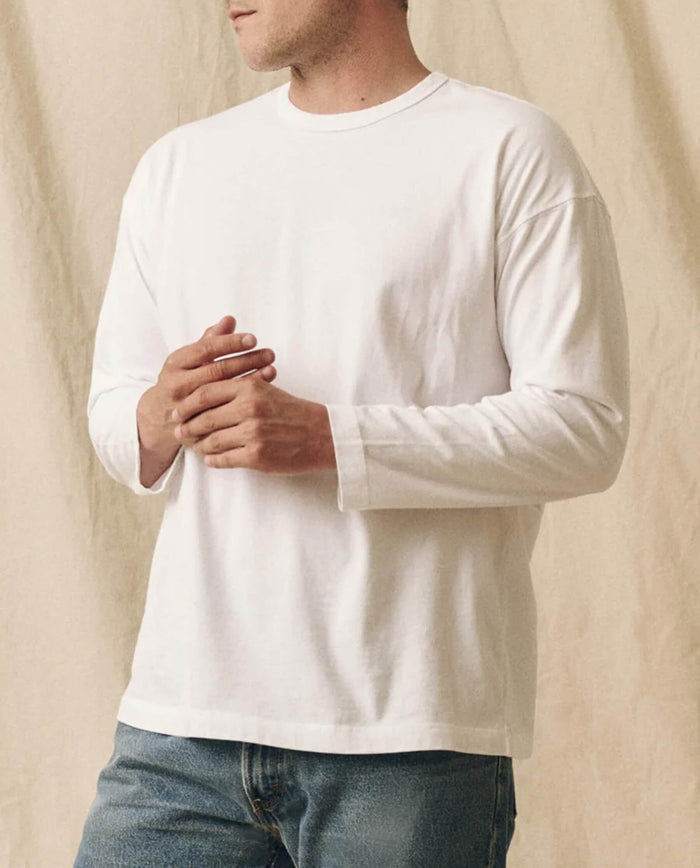 A person wears THE GREAT. FALL 23 MEN The Men's Pure Knits Long Sleeve Boxy Crew in Solid -- True White, made from 100% cotton, standing against a beige fabric backdrop with hands loosely clasped and facing slightly sideways.