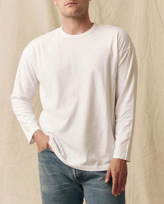 A person wearing THE GREAT. FALL 23 MEN The Men's Pure Knits Long Sleeve Boxy Crew in True White and blue jeans stands against a beige fabric background with one hand in their pocket.