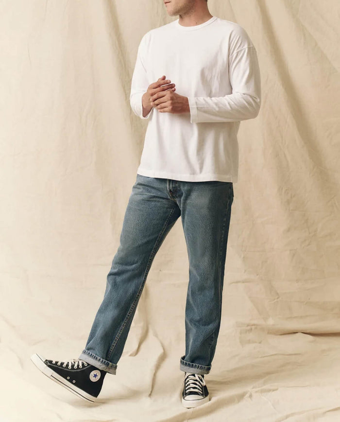 A person wearing THE GREAT. FALL 23 MEN The Men's Pure Knits Long Sleeve Boxy Crew in True White, blue jeans, and black Converse sneakers stands before a beige fabric backdrop.