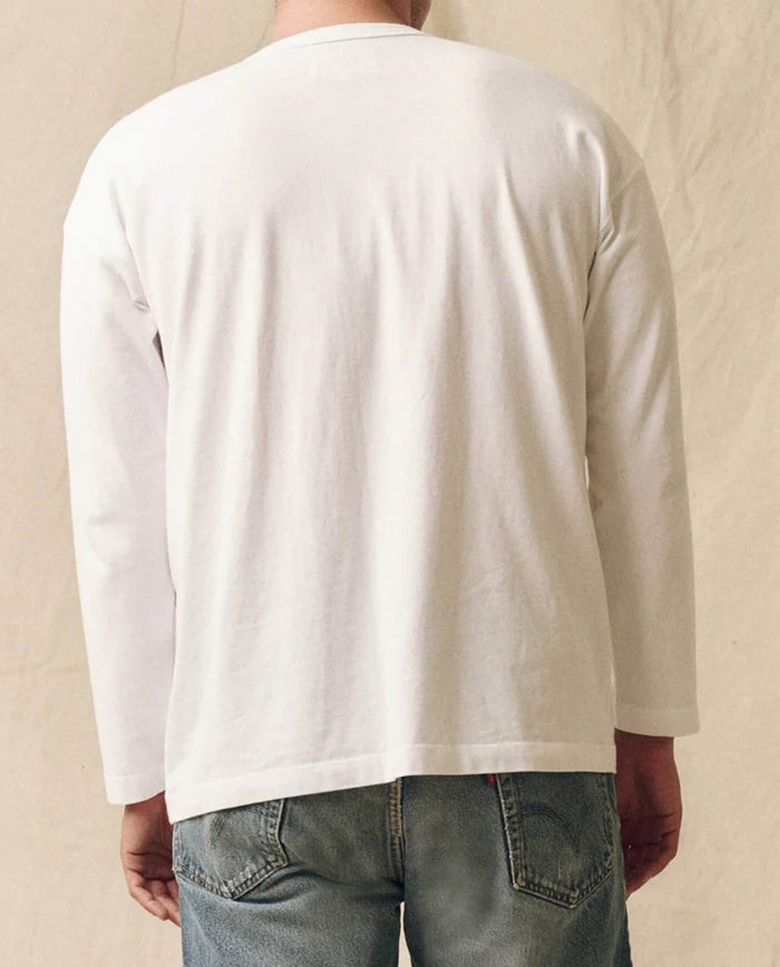 A person is seen from the back against a beige background, wearing THE GREAT. FALL 23 MEN - The Men's Pure Knits Long Sleeve Boxy Crew in Solid True White (100% cotton) paired with blue jeans.