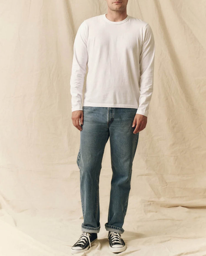 A person stands against a beige fabric backdrop, wearing THE GREAT. FALL 23 MEN The Men's Pure Knits Long Sleeve Slim Crew in True White, paired with blue jeans and black sneakers.