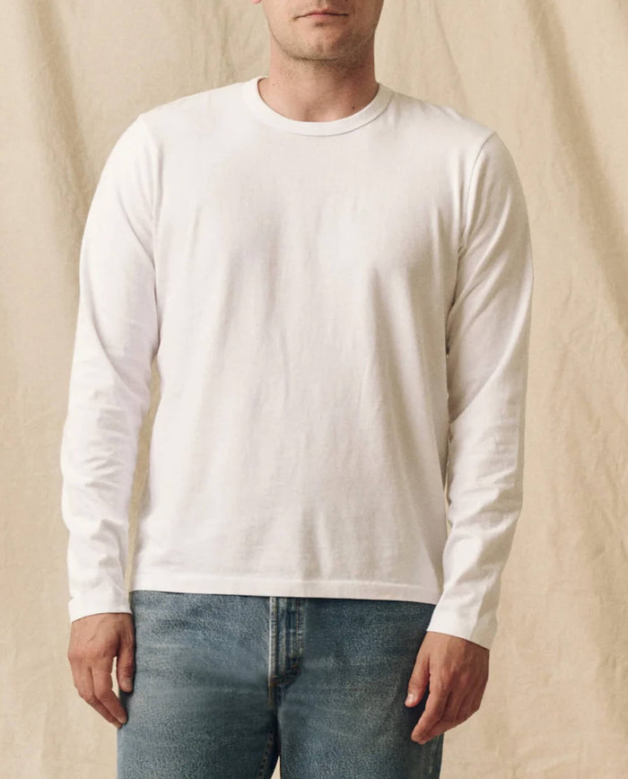 A person wears THE GREAT. FALL 23 MEN’s The Men’s Pure Knits Long Sleeve Slim Crew in True White with blue jeans, standing against a beige fabric background.