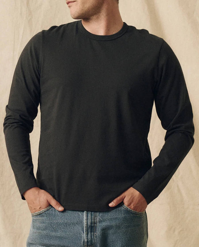 A person wears THE GREAT. FALL 23 MEN The Men's Pure Knits Long Sleeve Slim Crew in Solid -- Almost Black, made from heavy-weight fabric, with blue jeans and hands in pockets, standing against a beige background.