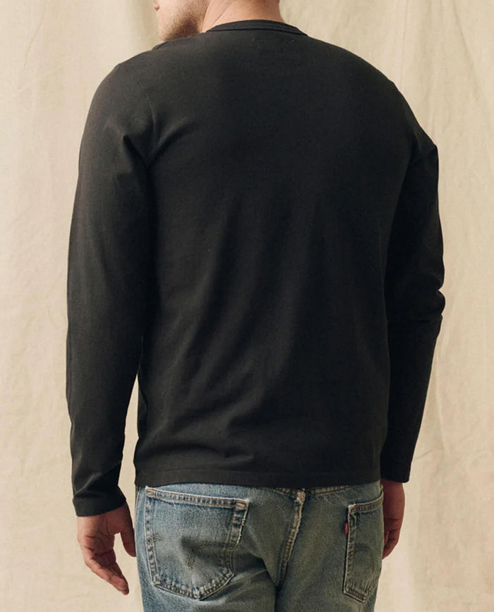 A person wearing THE GREAT. FALL 23 MEN The Men's Pure Knits Long Sleeve Slim Crew in Solid -- Almost Black stands facing away from the camera against a neutral background, paired with blue jeans.