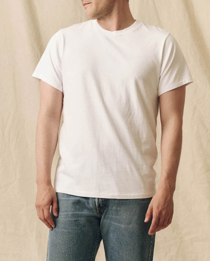 A person wears THE GREAT. FALL 23 MEN The Men's Pure Knits Slim Tee in True White with blue jeans, standing against a beige fabric background.