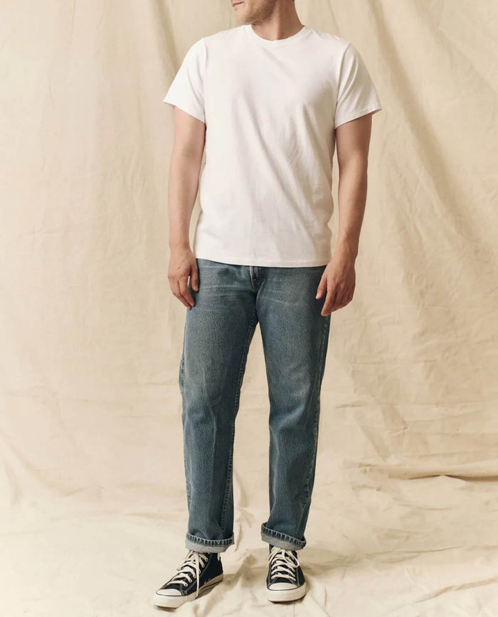 A person stands against a beige backdrop wearing THE GREAT. FALL 23 MEN's The Men's Pure Knits Slim Tee in True White, blue jeans with rolled cuffs, and black-and-white sneakers.