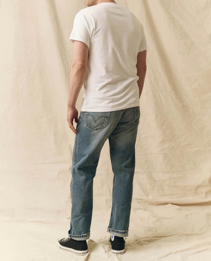 A person wearing THE GREAT. FALL 23 MEN The Men's Pure Knits Slim Tee in True White, along with blue jeans and black sneakers, stands facing away against a beige fabric backdrop.