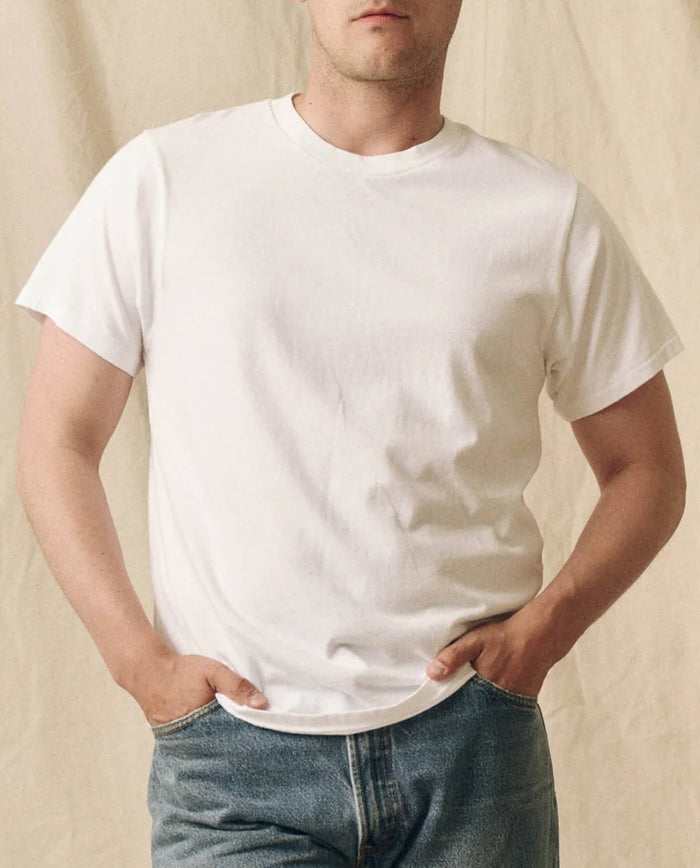 A person wears THE GREAT. FALL 23 MEN The Men's Pure Knits Slim Tee in True White and blue jeans, standing against a beige background with hands in pockets.