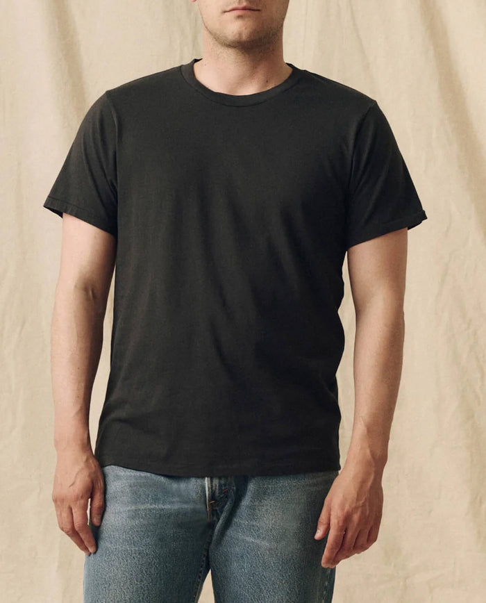 A person wearing THE GREAT. FALL 23 MEN The Men's Pure Knits Slim Tee in Solid -- Almost Black and blue jeans stands before a beige fabric background.