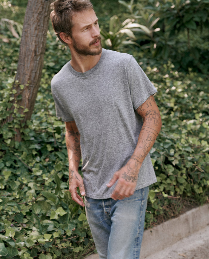 A tattooed man stands outdoors against a backdrop of lush green foliage, wearing a vintage-inspired Heather Grey Men's Slim Tee from THE GREAT. MAN and blue jeans.