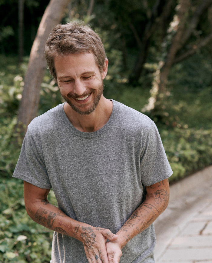 A man with short hair and a beard, wearing The Men's Slim Tee in Heather Grey from THE GREAT. MAN, smiles while looking down. He is outdoors, surrounded by greenery and trees. His arms are covered in tattoos.