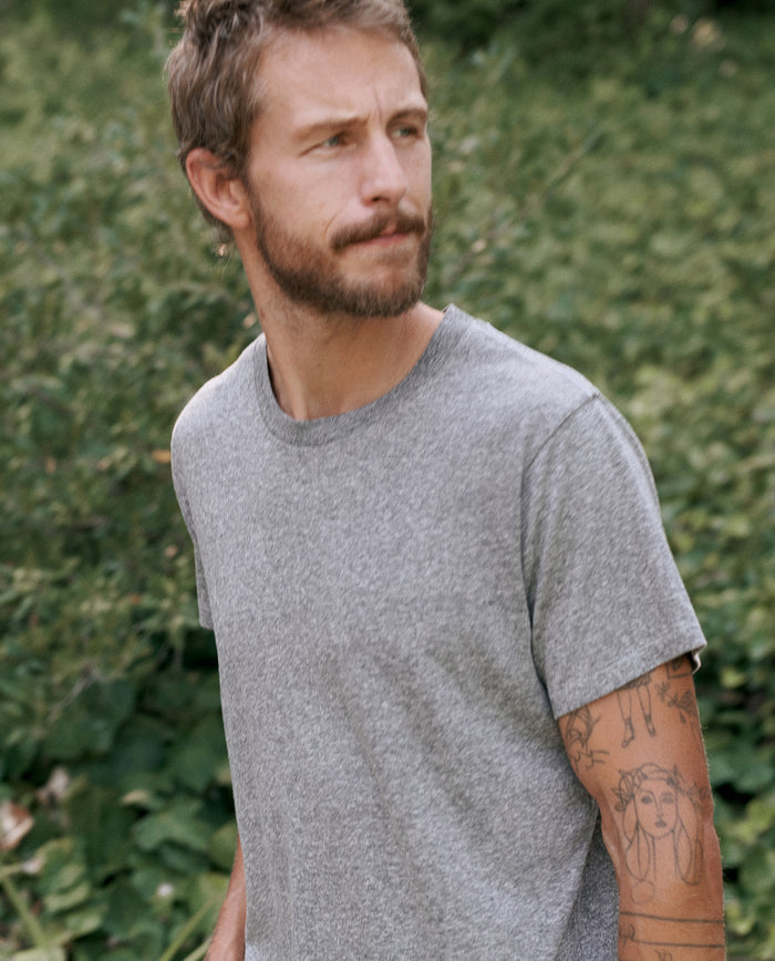 A man with short hair and a beard is wearing The Men's Slim Tee in Heather Grey by THE GREAT. MAN, standing outdoors with greenery in the background. His right arm has visible tattoos.