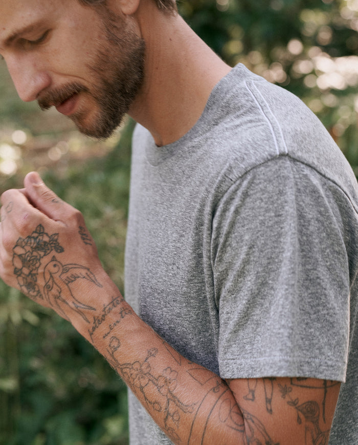 A man with tattoos on his arms, wearing The Men's Slim Tee in Heather Grey by THE GREAT. MAN, stands outdoors.