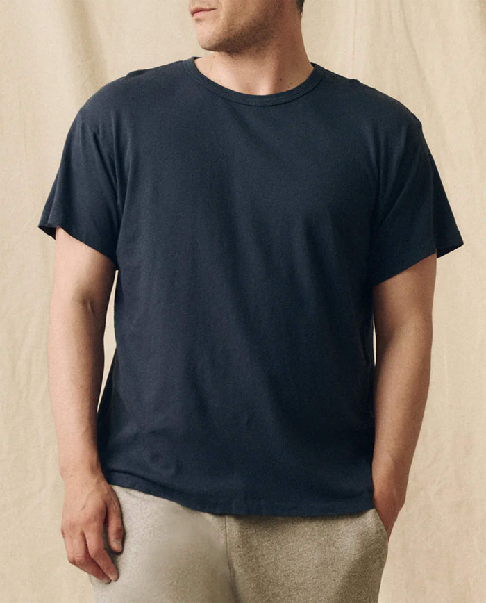 A man wears THE GREAT. FALL 23 MEN The Men's Slim Tee in True Navy with light-colored pants, standing before a beige fabric backdrop.