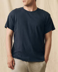 A man wears THE GREAT. FALL 23 MEN The Men's Slim Tee in True Navy with light-colored pants, standing before a beige fabric backdrop.