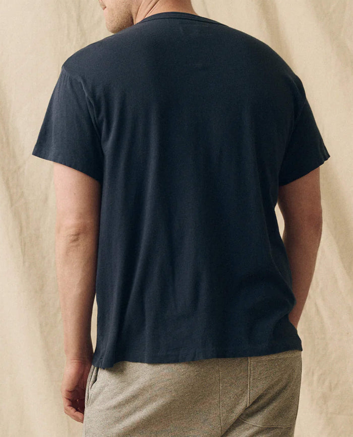 A man stands with his back to the camera, wearing THE GREAT. FALL 23 MEN The Men's Slim Tee in True Navy and light-colored shorts, set against a neutral fabric backdrop.