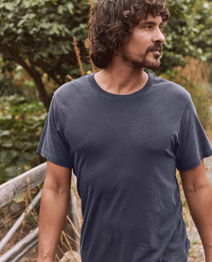 A man with curly hair and a beard is walking outside, wearing The Men's Slim Tee in washed navy from THE GREAT. SP22 D2 MENS. Green foliage is visible in the background.
