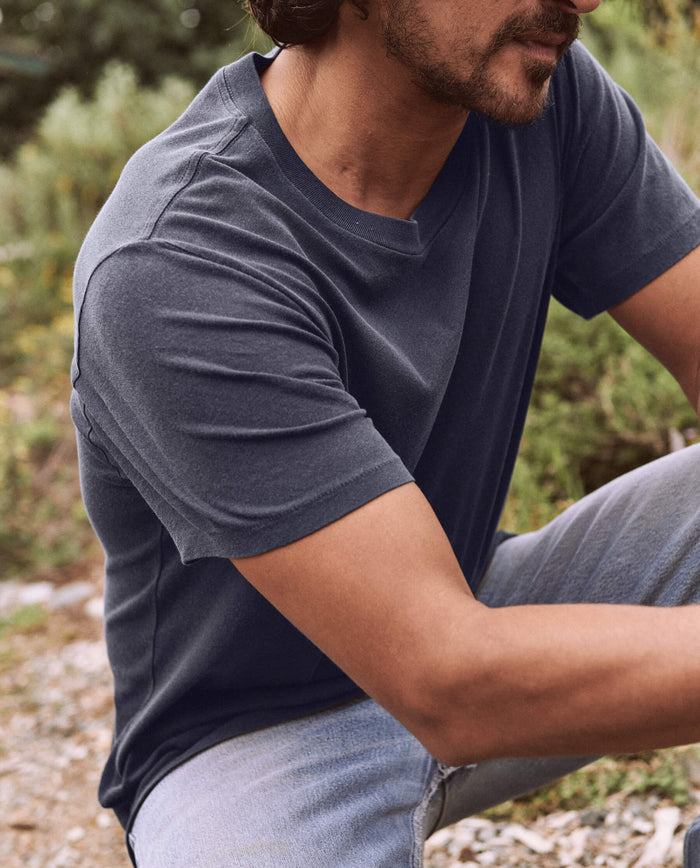 A person wearing The Men's Slim Tee in Washed Navy by THE GREAT. SP22 D2 MENS and blue jeans is crouching outdoors in a natural setting with greenery in the background.