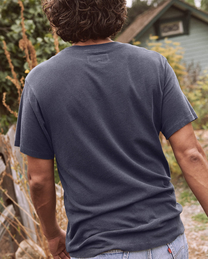 A person with wavy hair, wearing The Men's Slim Tee in Washed Navy by THE GREAT. SP22 D2 MENS and jeans, is standing outdoors with their back facing the camera. In the background, there is foliage and a house.