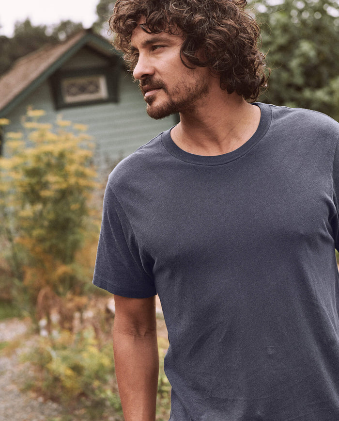 A man with curly hair and a beard stands outdoors wearing The Men's Slim Tee in Washed Navy by THE GREAT. SP22 D2 MENS, with a small green house and foliage in the background.