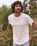 A man with curly hair and a beard stands outdoors, wearing The Men's Slim Tee in Washed White by THE GREAT. HOL 21 MENS and light blue jeans, with greenery in the background.
