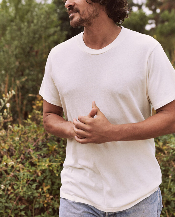 A man wearing The Men's Slim Tee in Washed White by THE GREAT. HOL 21 MENS stands outdoors with hands clasped, greenery in the background, paired with jeans.