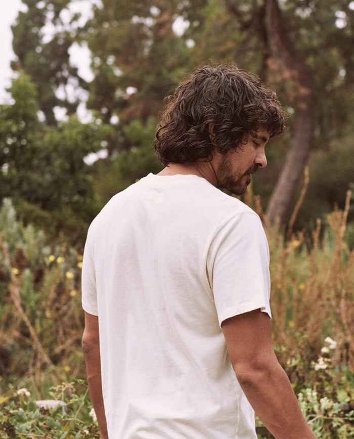 A man with curly hair wearing The Men's Slim Tee in Washed White by THE GREAT. HOL 21 MENS stands in a natural outdoor setting with trees and foliage in the background.