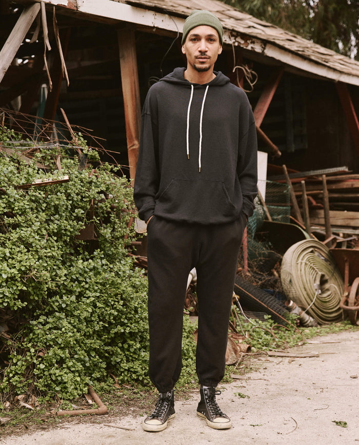 A man wearing The Men's Stadium Sweatpant in Almost Black from THE GREAT. HOL 21 MENS stands with his hands in his pockets, donning vintage athleticwear in front of a rustic shed adorned with greenery, various debris, and coiled wire.
