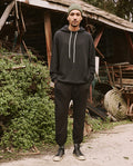 A man wearing The Men's Stadium Sweatpant in Almost Black from THE GREAT. HOL 21 MENS stands with his hands in his pockets, donning vintage athleticwear in front of a rustic shed adorned with greenery, various debris, and coiled wire.