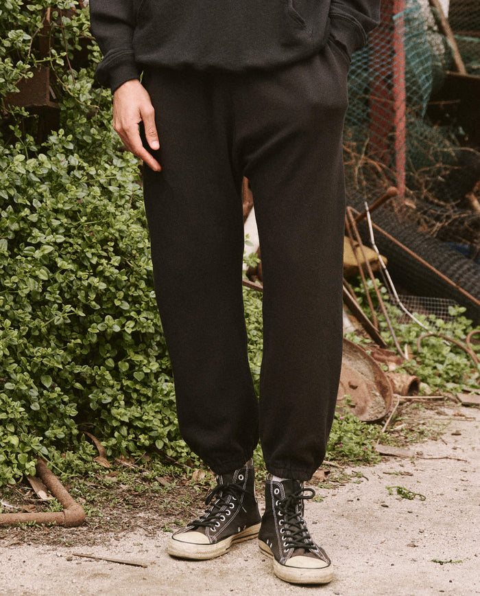 A person wearing The Men's Stadium Sweatpant in Almost Black by THE GREAT. HOL 21 MENS and black high-top sneakers, standing beside a patch of greenery.
