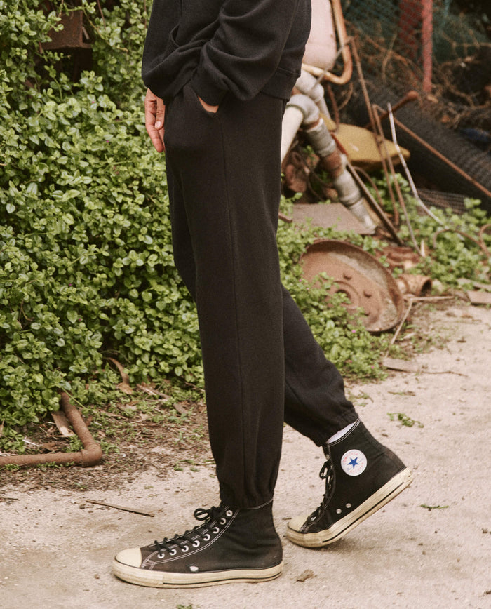Person wearing The Men's Stadium Sweatpant in Almost Black from THE GREAT. HOL 21 MENS along with black high-top sneakers with white soles, standing on a path next to some greenery and rusty debris.
