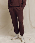 Person standing against a beige backdrop, wearing THE GREAT. FALL 24 MEN's Toasted Walnut Men's Stadium Sweatpants, a matching sweatshirt, and white sneakers, with hands in pockets.