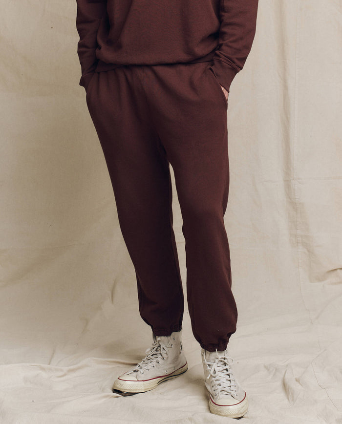 Person standing against a beige backdrop, wearing THE GREAT. FALL 24 MEN's Toasted Walnut Men's Stadium Sweatpants, a matching sweatshirt, and white sneakers, with hands in pockets.