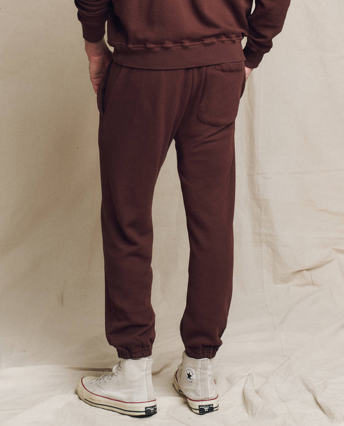 A person is seen from the back, dressed in The Men's Stadium Sweatpant in Toasted Walnut by THE GREAT. FALL 24 MEN, and white high-top sneakers, standing on a beige fabric background. The look exudes vintage athleticwear charm with these classic sweatpants, seamlessly blending old-school style with modern flair.