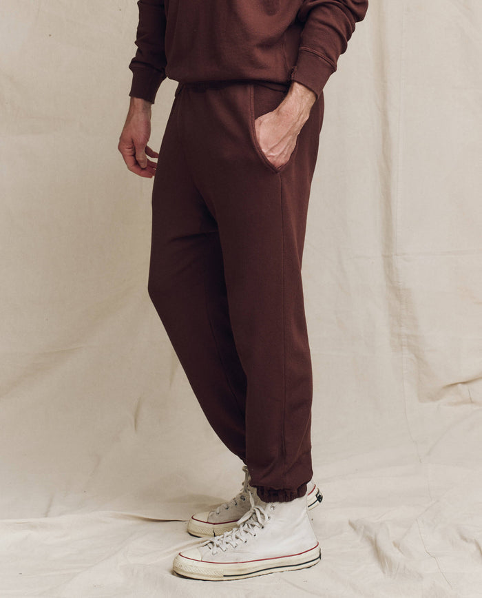 A person wearing "The Men's Stadium Sweatpant" in Toasted Walnut by THE GREAT. FALL 24 MEN, reminiscent of vintage athleticwear, and white high-top sneakers stands against a cream-colored backdrop, with one hand in their pocket.