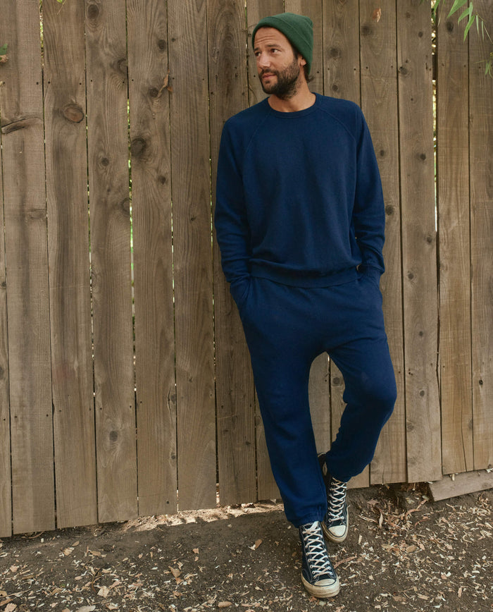 A man in a navy blue outfit stands against a wooden fence, his hands in his pockets and his green beanie adding a pop of color. He completes his look with black and white sneakers and The Men's Stadium Sweatpant by THE GREAT. HOL 22 MEN, giving him an effortlessly cool vibe.