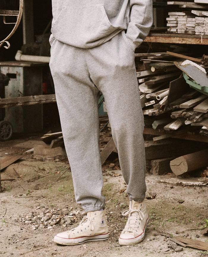 A person standing on a dirt ground, scattered with wooden planks and metal pieces in the background, is wearing The Men's Stadium Sweatpant - Varsity Grey by THE GREAT. HOL 21 MENS and white high-top sneakers.